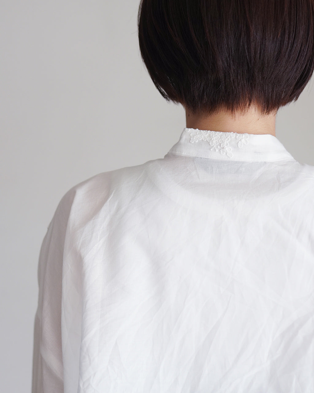mizuiro indの白シャツ、後ろはスタンドカラーに小さなレース風の刺繍ディテールが映える仕様。首元のアップショットで素材のしわ感と清潔感を伝える1枚。