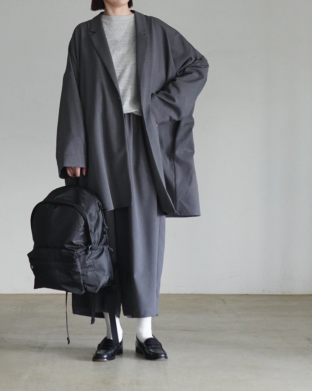 グレー系のwide tailored shirt JKを着用した女性の全身ショット。ロング丈の oversized ジャケットが垂れ下がり、背面と前方のバランスが分かる構図。白いソックスと黒のローファーを合わせ、ミニマルな印象。