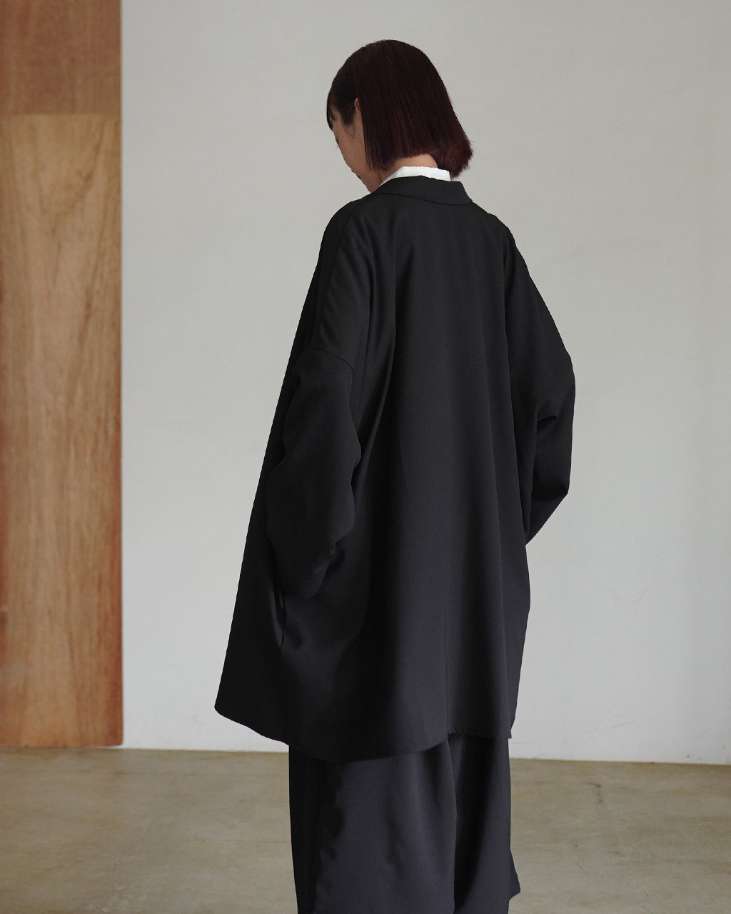 背面から見た黒のジャケット。ゆったりとしたビッグシルエットで、長めの丈と落ち着いた印象の佇まいが特徴的です。