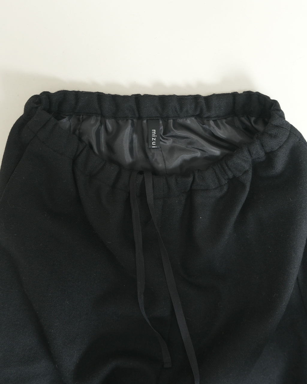 ウール混のリラックスシルエットのパンツ裾まで覆うブラックカラー、ウエストは絞り紐付きのイージー仕様。ブランドタグが内側ウエストに見えるディテール画像。