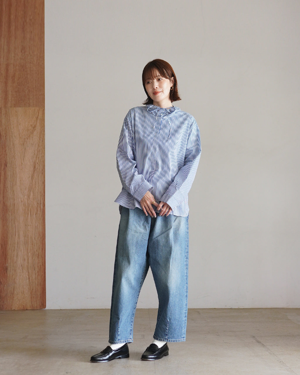 青と白のストライプ柄シャツを着た女性の全身写真。ゆるめのシルエットで裾が広がり、ダメージデニム風のパンツと黒のローファーを合わせたカジュアルなコーディネート