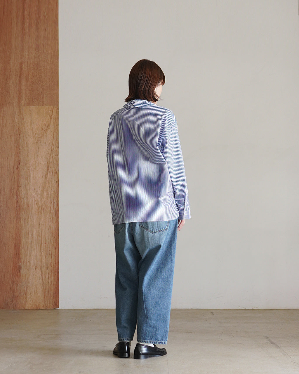 青と白のストライプ柄シャツを後ろ姿で着用した女性。ゆったりとしたシルエットで裾は腰周りに落ち、パンツはデニム。背中の折り返しが見えるデザイン。
