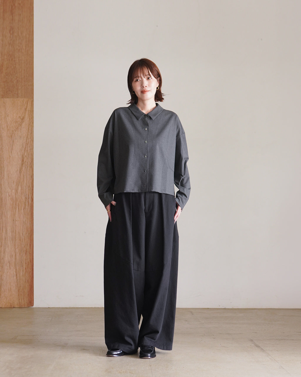 mizuiro indのT/Rワイドシャツを着用したモデルが正面に立つシーン。グレー系の長袖シャツで、ゆったりとしたシルエットと裾がやや短め。黒系パンツと合わせたコーディネート。