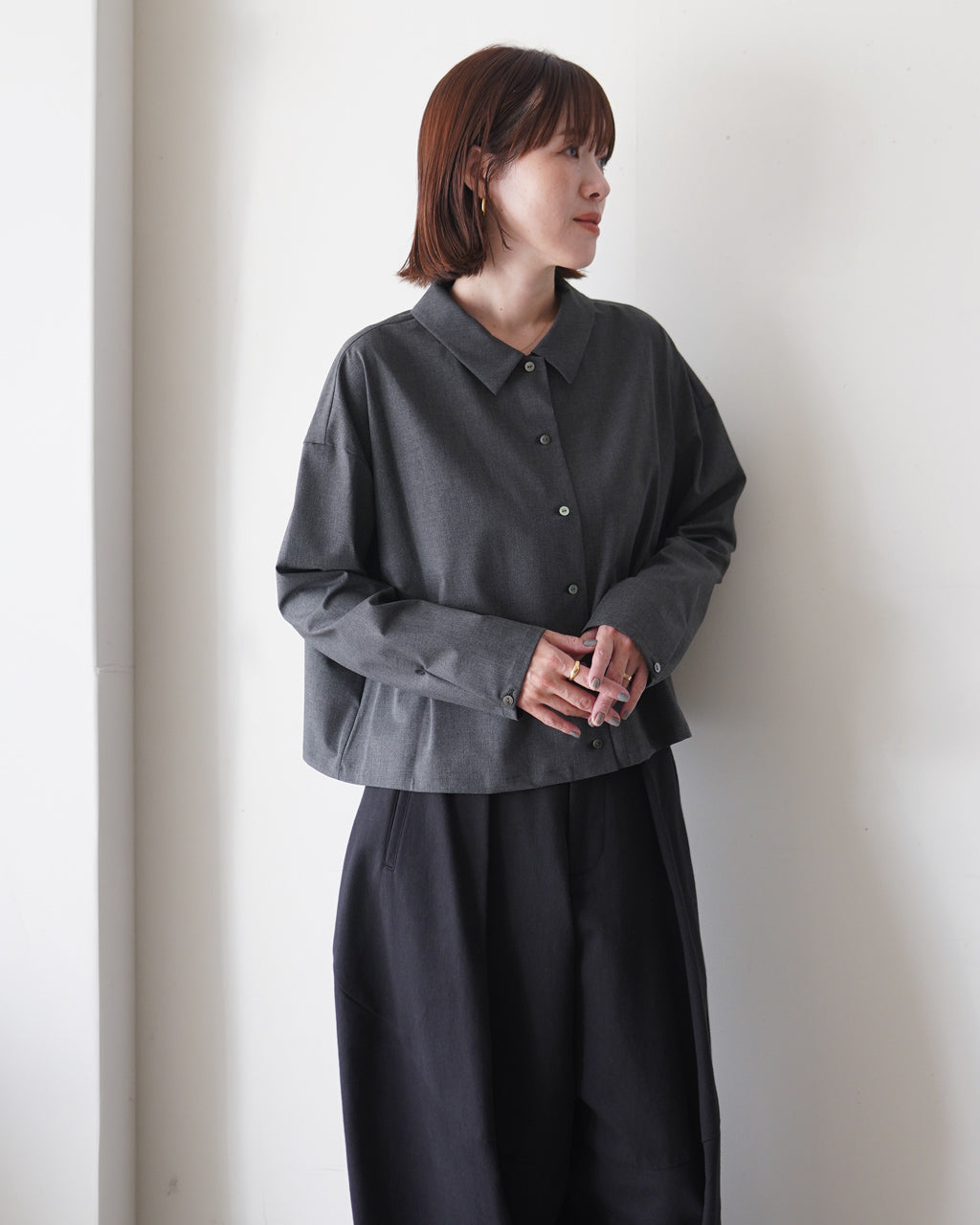 濃いグレーの長袖シャツを着た女性が壁際で横向きに立つ様子。ゆったりとしたシルエットで前ボタンが中央に並ぶデザイン。