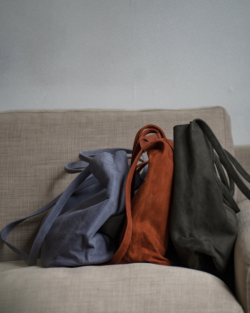ソファに並ぶ3カラーのモルフェ3WAYミディウムトート。オイルスエード素材の柔らかな質感と、バックスタイルの持ち手が見える横置きショット