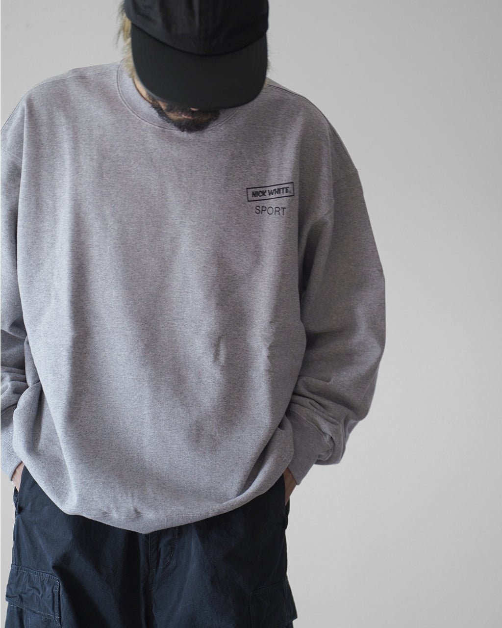 グレーのクルーネックスウェットを着用した男性の上半身。胸元に小さくロゴが入ったシンプルなデザインで、帽子と合わせたカジュアルな着こなしが印象的です。