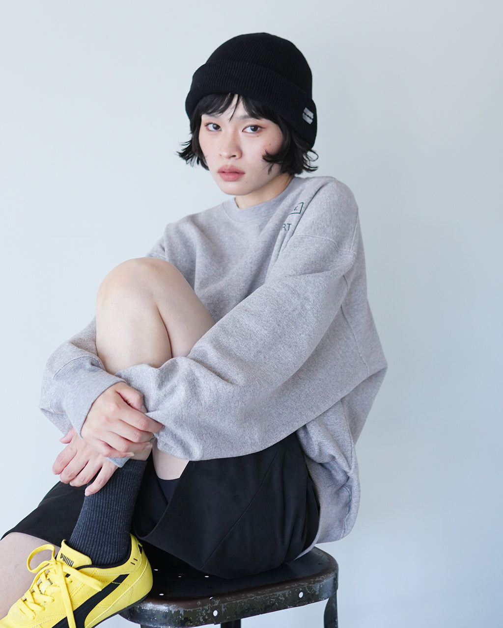 灰色クルーネックスウェットを着た女性が座り、膝を抱えたポーズで撮影。ゆったりしたシルエットと厚みのある生地感が伝わる前面ショット。