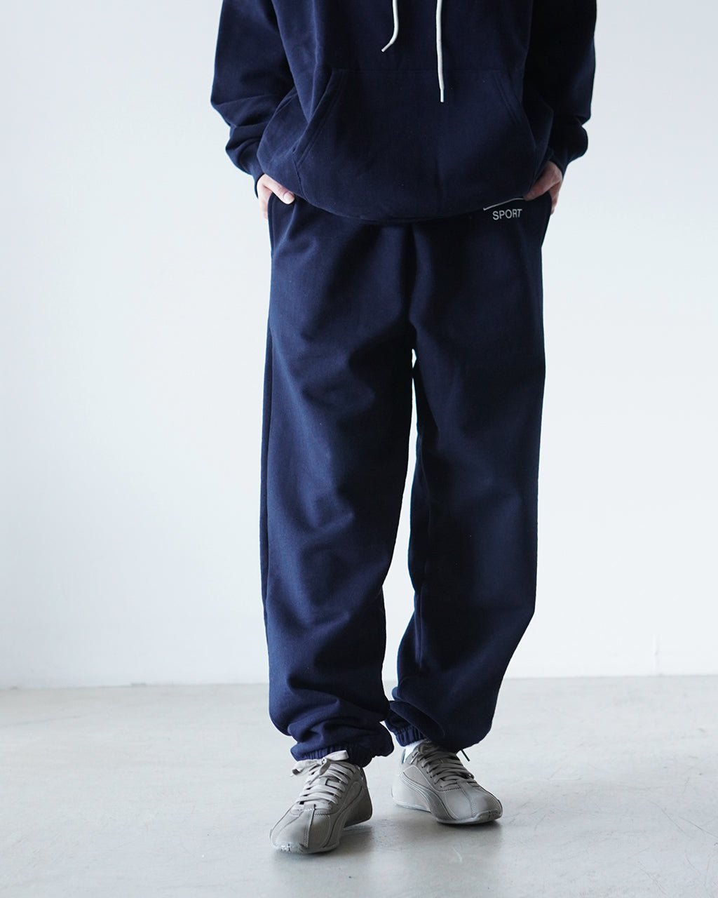 ネイビーのスウェットパンツと同色のパーカーのセットアップ風ルック。腰まわりのポケットとロゴ近くの“SPORT”刺繍が見える裾までゆとりのあるシルエットの着用イメージ