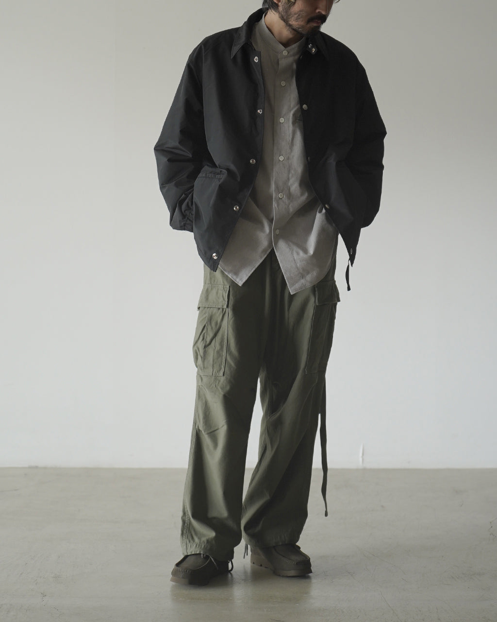 ナイジェル・ケーボンのARMY CARGO PANTを着用した男性の全身写真。黒のジャケットの下にグレーのシャツを合わせ、カーキのカーゴパンツが強調される正面全身ショット。