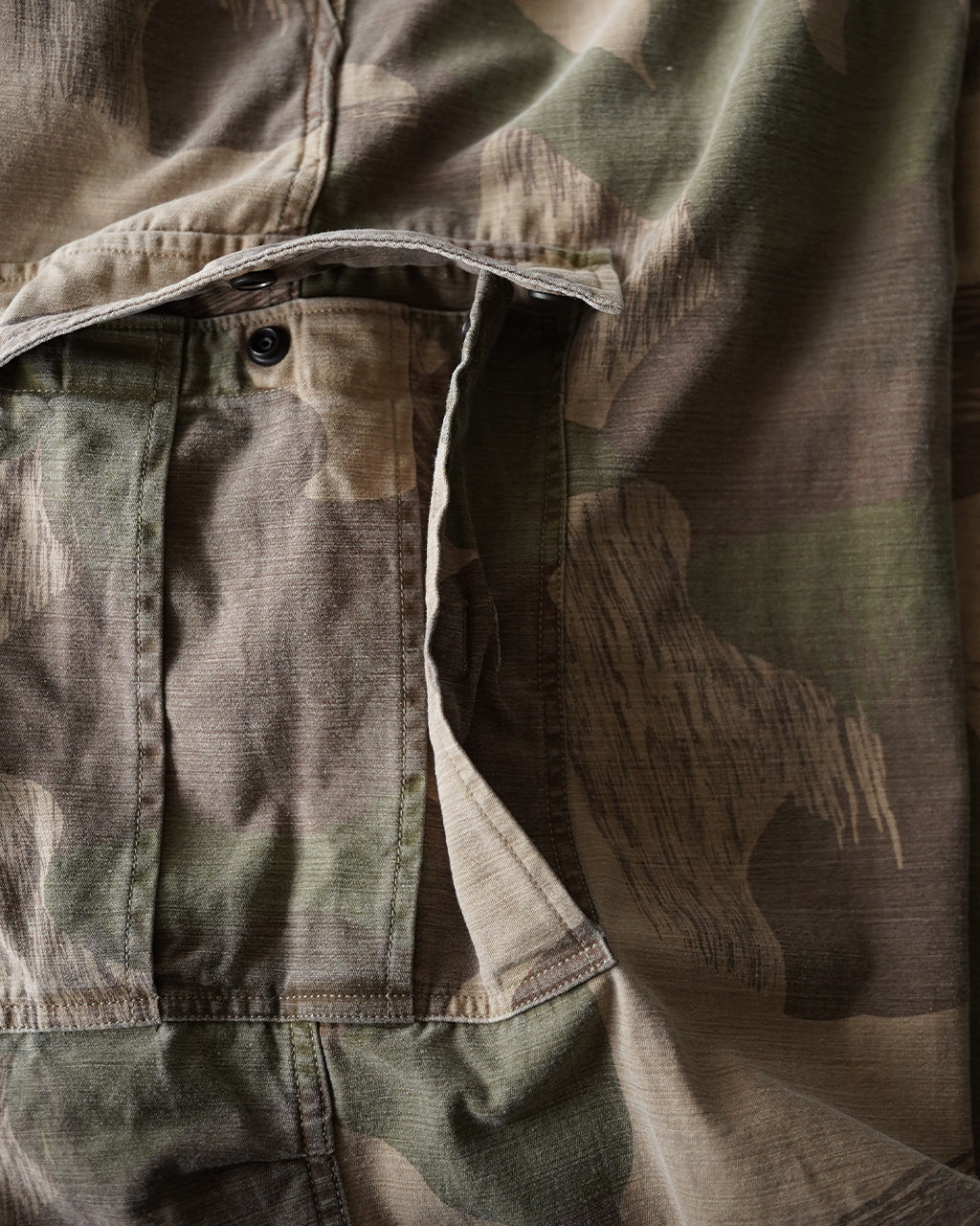 ナイジェル・ケーボンのARMY CARGO PANT FADE CAMOの前後を示す軍用カーゴパンツ。迷彩柄の大きなポケットと前開きディテールが特徴的で、素材はコットンの質感を近くで見えるリベットと縫製で表現。