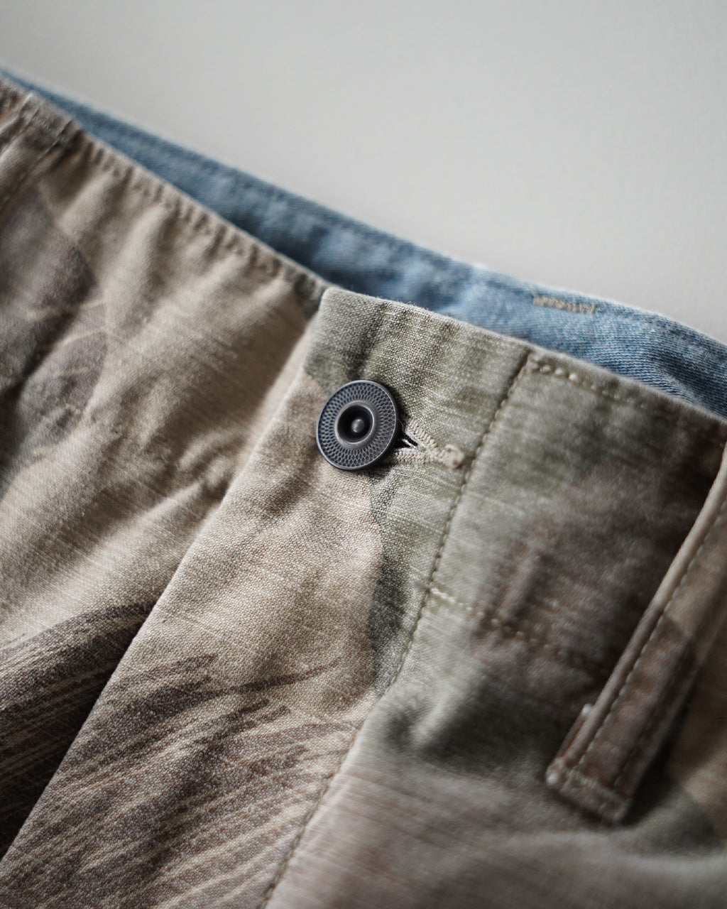 カーキとベージュの迷彩生地に近い色味のARMY CARGO PANTのウエストまわりをアップ。ボタンと布地の質感が強調されたディテールショット。