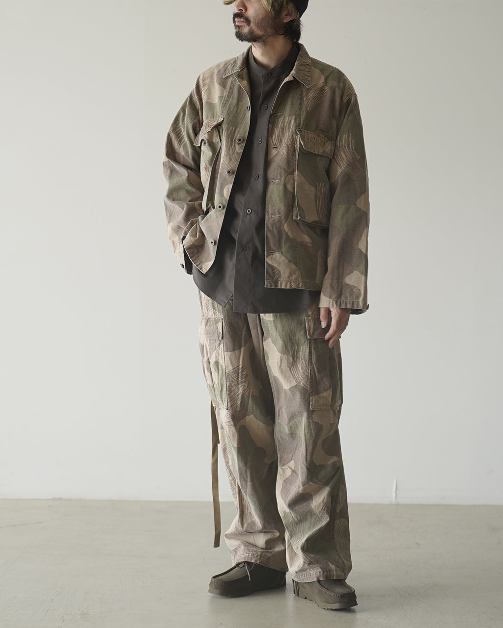 FIELD JACKET FADE CAMOを着用した男性モデルの全身画像。迷彩柄のジャケットと同色系のパンツを組み合わせ、ゆとりのあるシルエットが特徴。