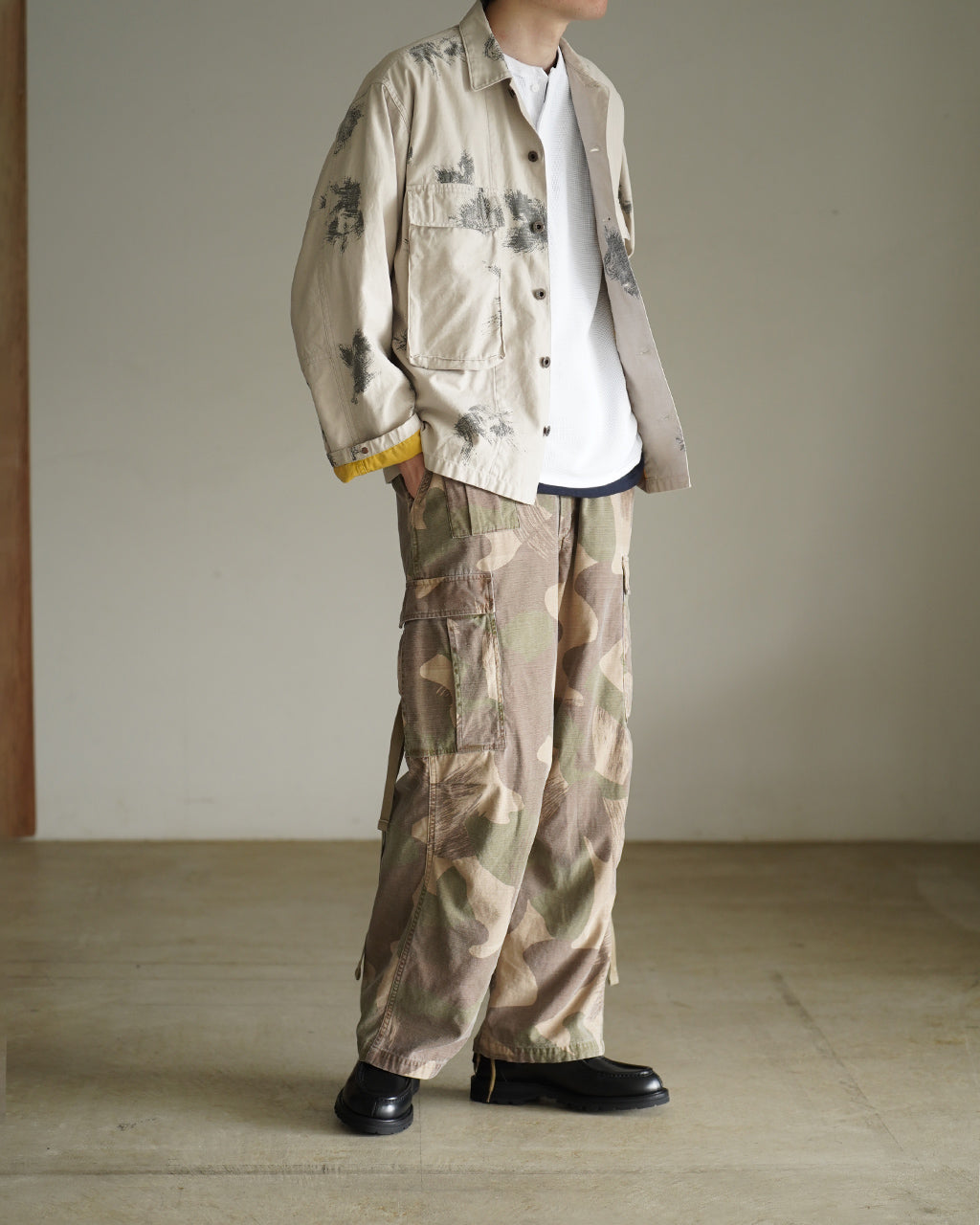 着用イメージのFIELD JACKET FADE CAMOを横から見た写真。ベージュ系の迷彩ジャケットとカーゴパンツの組み合わせ。インナーは白いニットで、裾や袖のシルエットが分かる。