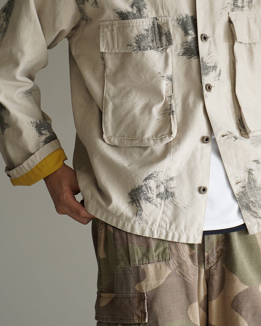 FIELD JACKET FADE CAMOの正面アップ。淡いベージュ系のジャケットに黒系の迷彩プリントが散らばり、胸ポケットと前ボタンが視認できるクローズショット。