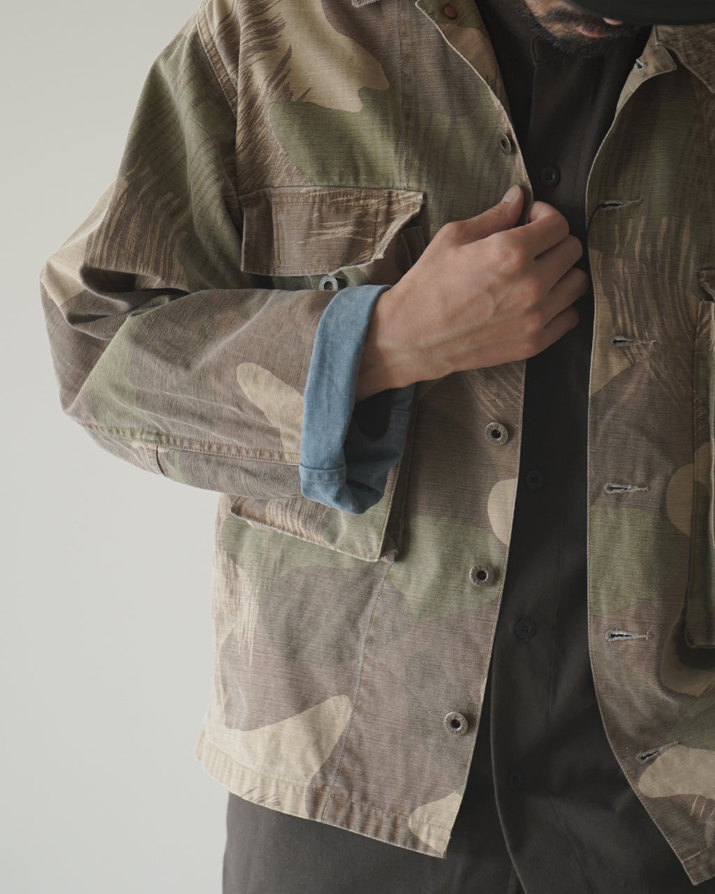 FIELD JACKET FADE CAMOのカモフラージュ柄ジャケットを腰近くまで着用したアップショット。前面ポケットやボタン、袖口のディテールが見える。裏地はなしの一重仕立てで、生地の質感と色味の風合いが伝わるショット。