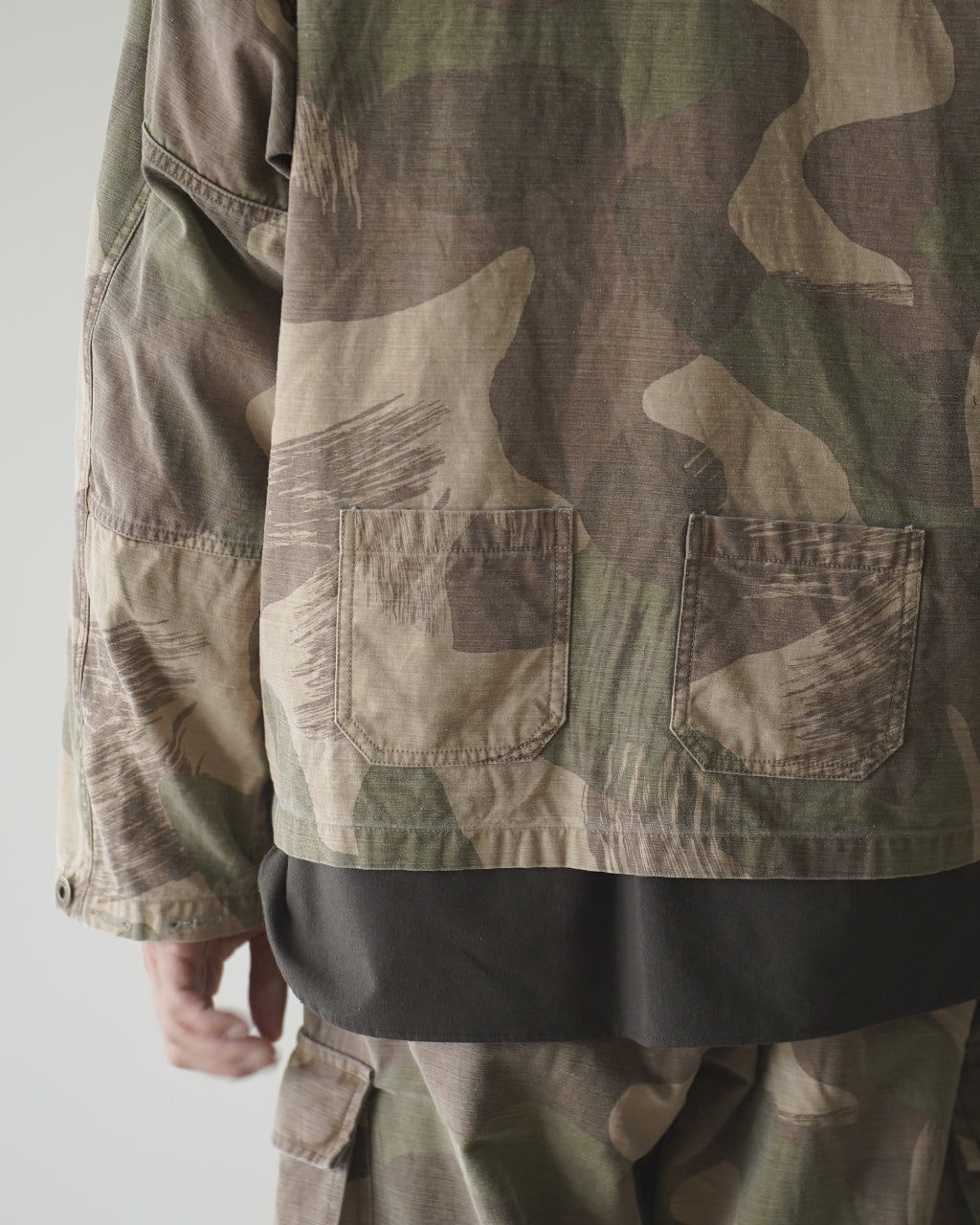 FIELD JACKET FADE CAMOの背面と裾が見えるカモフラージュ柄ジャケット。ウエスト周りのポケットと下部のブラックカラーの裾がアクセントになり、袖口にはボタン留めが見える。