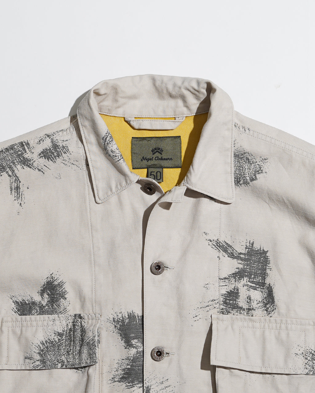フェードカラーのカモフラージュ柄が施された FIELD JACKET のアップショット。淡いベージュ系ボディにダークグレーのプリント風模様が散らばるデザインを正面から捉えた画像。