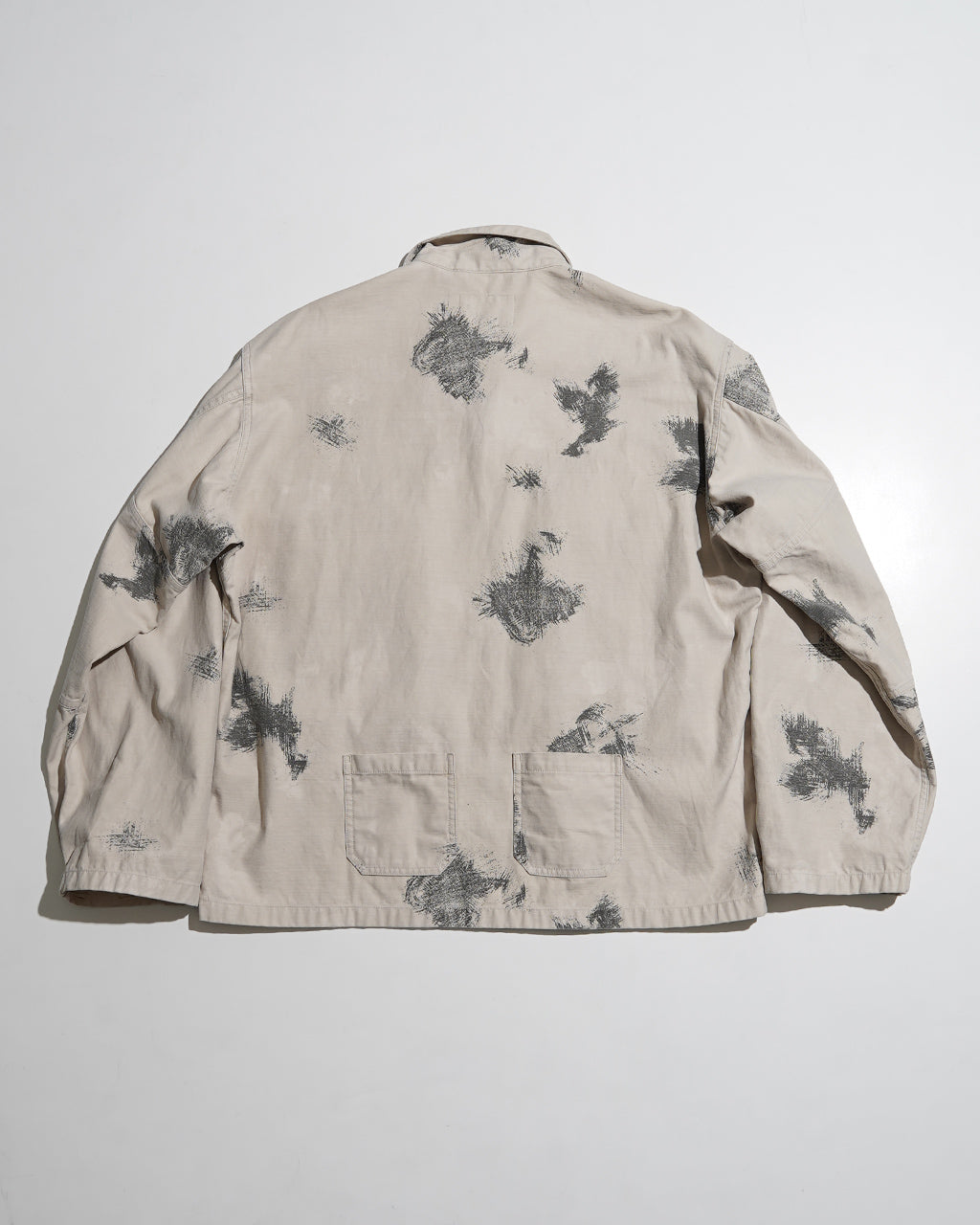 裏返しのような淡いベージュ地に、黒系の飛び柄プリントが散らばるFIELD JACKETの背面ショット。裾近くにはポケットが二つ並び、ワーク感とミリタリーテイストを両立させたデザインが分かる。
