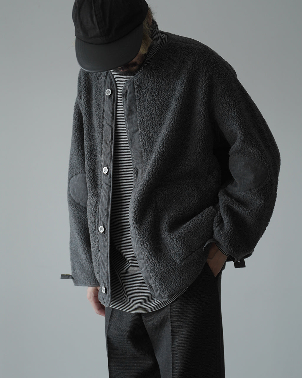 M-65 FISH TAIL COAT LINERの着用イメージ。黒系のボアフリース素材が全体を覆い、前立てと袖口に別布パネルが見える。帽子をかぶりストライプのインナーと組み合わせたスタイル。