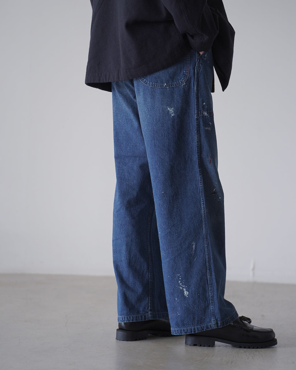 デニム生地のゆるやかなワイドストレートパンツ、後ろポケット横の縫製と裾のディテールが特徴。深いインディゴカラーに洗い加工が施され、ペイント風の斑点がアクセントとして現れているレッグショット。