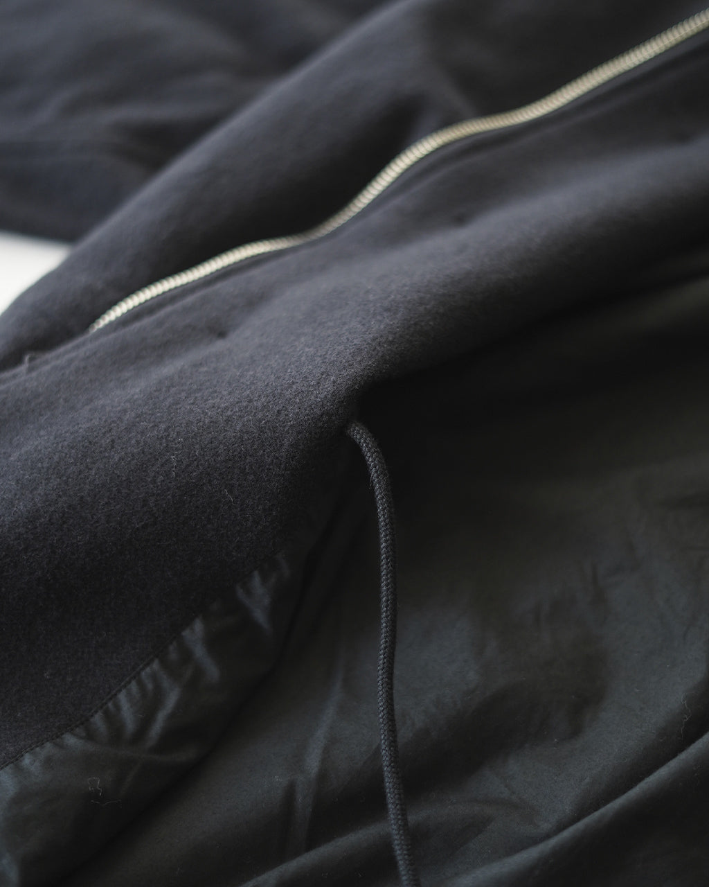 MANTEAU ASSATのチャコール色生地に沿ったジッパーとドローコードをアップで捉えた拡大ショット。表生地の質感と滑らかな裏地の組み合わせが分かるディテール画像