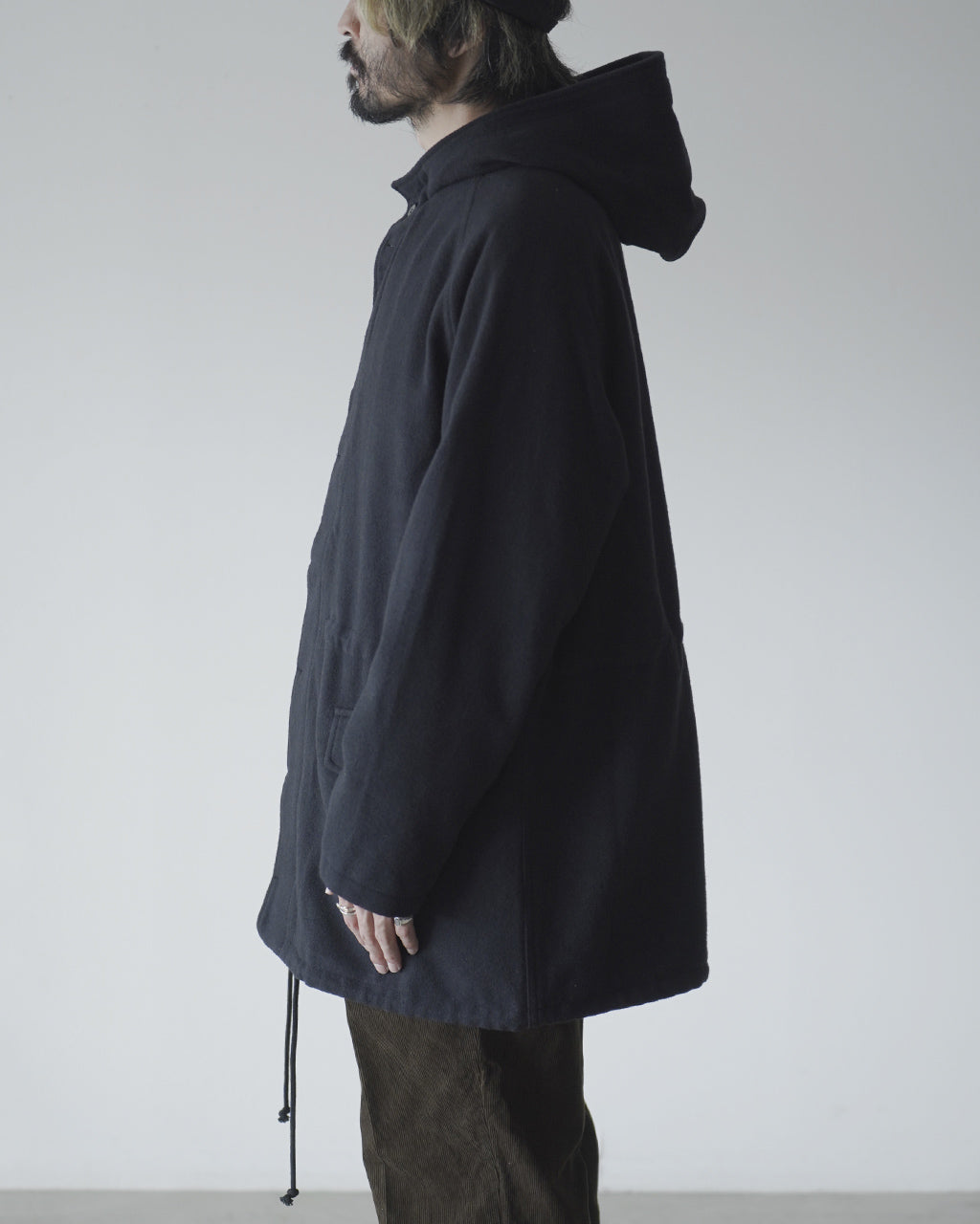 黒のフード付きジップアップコート前身頃が見える横姿。ゆったりとしたシルエットで裾にドローコードが垂れ、袖口はやや長めに感じられるデザインです。
