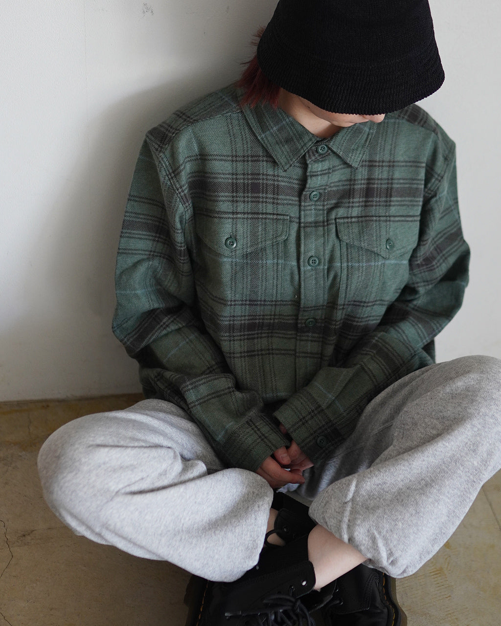 緑系のチェック柄シャツを着た人物が壁際に座って前方を見ていない。胸ポケット付きのフロントとクラシックなボタンダウンが特徴。