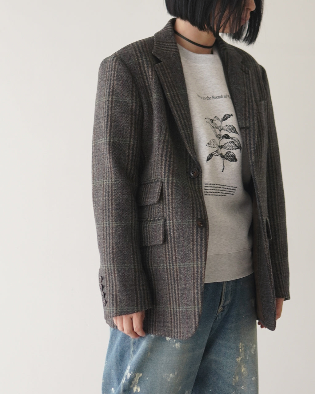 グレー系チェック柄のジャケットを着た人物の横からの写真。下にはライトグレーのスウェットとデニムパンツを合わせ、胸元の植物プリントが控えめに覗くカジュアルスタイル。