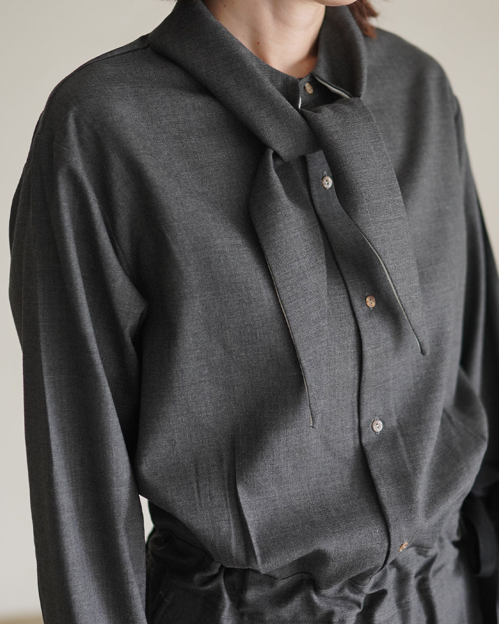 グレーのFine Worsted Jumpsuitのアップショット。首元の前結びリボンタイとシャツ風ボタンが際立つ布地の質感を捉えています。