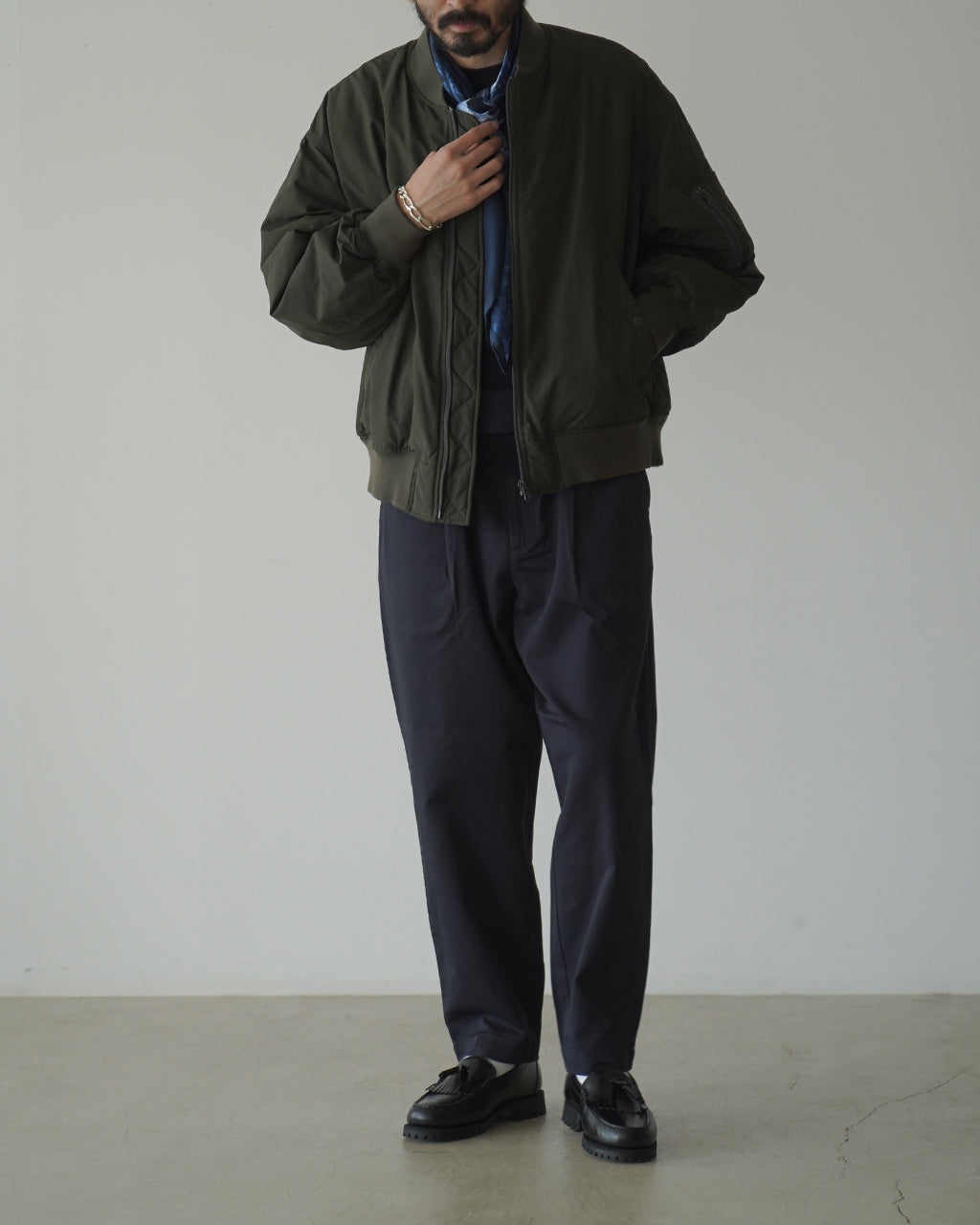 olive色のボンバージャケットを着用した男性が立ち、裾に向かって細身になるシルエットが特徴的なコーディネート。全体はシックな印象。