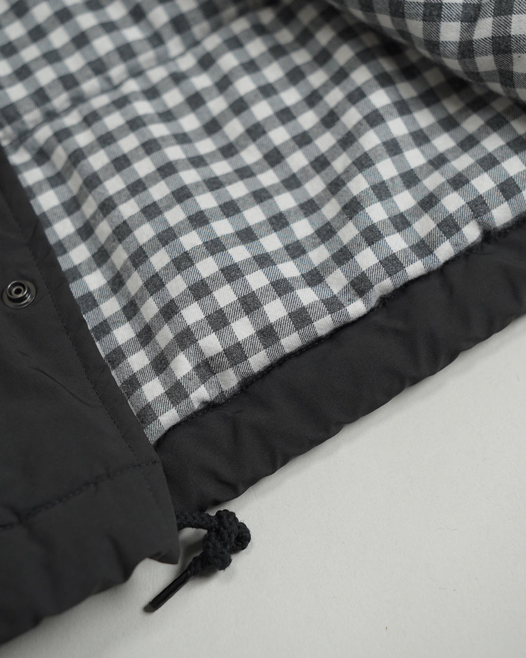 チェック柄の裏地が際立つ Weather Coach Jacket の裾とボタン周りのアップ。黒の縁取りと絞りコードが特徴的なディテール写真。