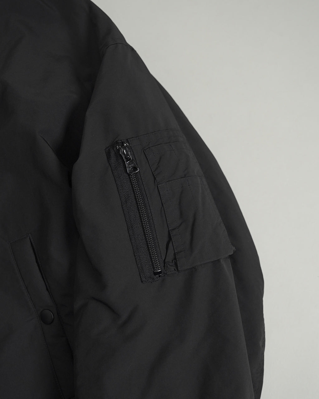 黒のWeather Flight Jacketの左腕ポケット横のジップ付きファスナーポーチをアップで捉えたディテールショット。耐候性のあるマットな表地とポケット縫い目のディテールが際立つ。落ち着いた雰囲気のジャケットの質感が伝わる一枚。
