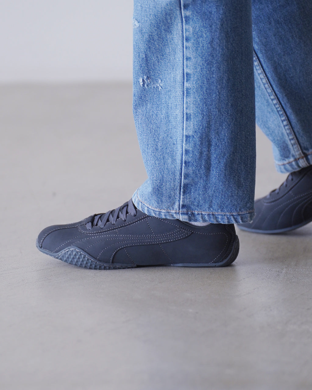 PUMAの紺色系スニーカーの横から覗く突起のないつくり。ヌバック風の表面と縫い目が映えるデザインで、ジーンズと合わせたカジュアルな足元を描写。