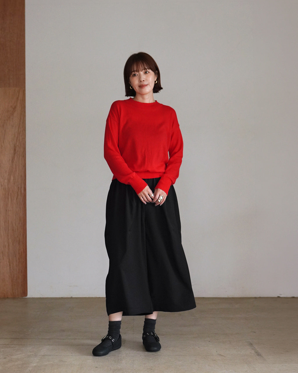 赤い長袖ニットを胸元まで見せた女性が黒のロングスカートを着用。室内のシンプルな背景で、カジュアルながらきちんとした印象のコーディネートを表現。