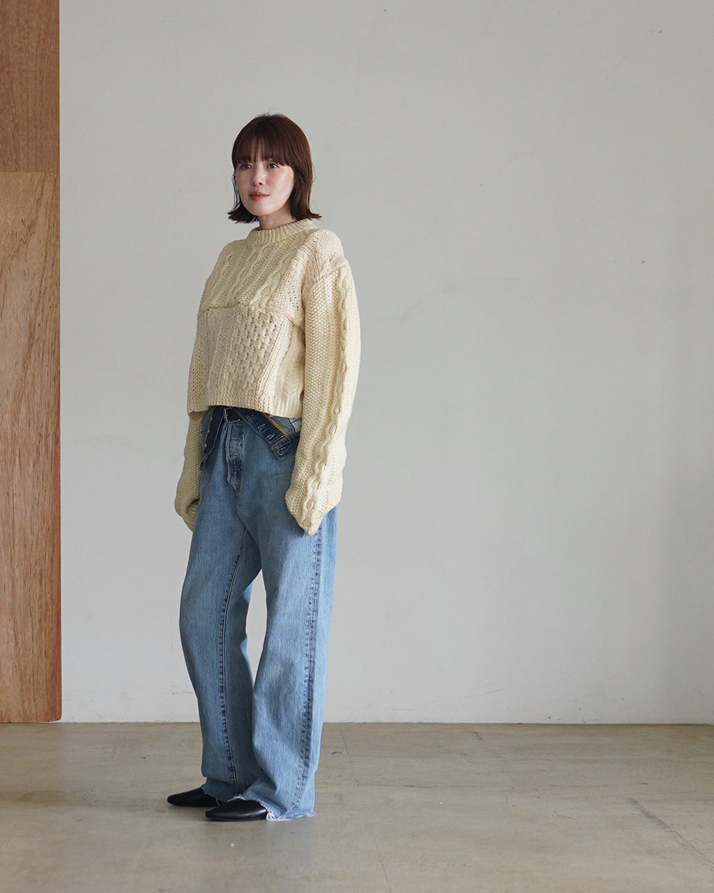オフホワイト系のケーブル編みニットを着た女性モデルの全身画像。肩から袖までボリュームのある長袖デザインが特徴。デニムパンツと黒い靴でカジュアルな着こなし。