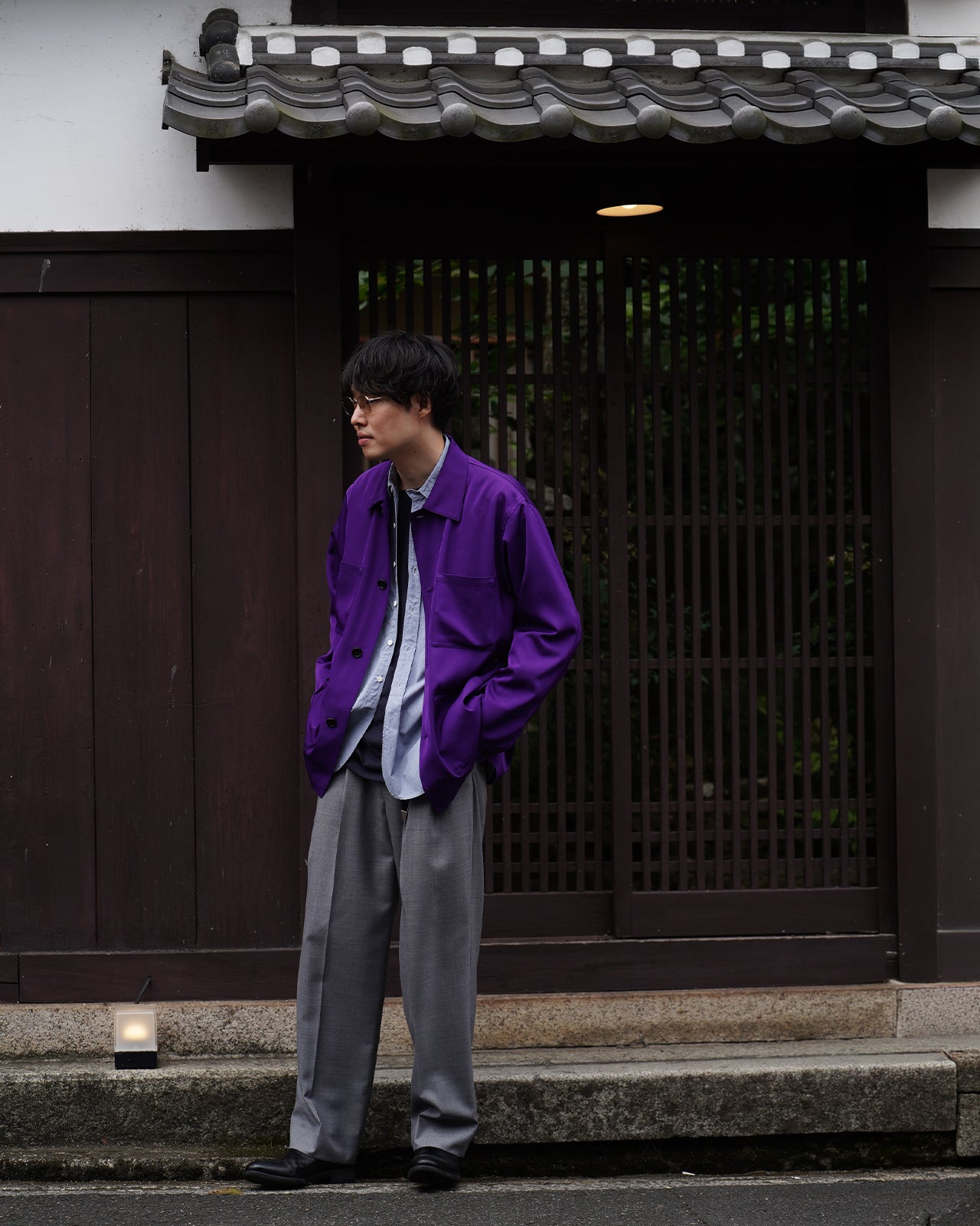 京都の門前に立つ男性が、紫のレインメーカーのジャケットを着て横向きにポケットへ手を入れている。落ち着いた色味と光沢のある生地感が特徴的な着用写真