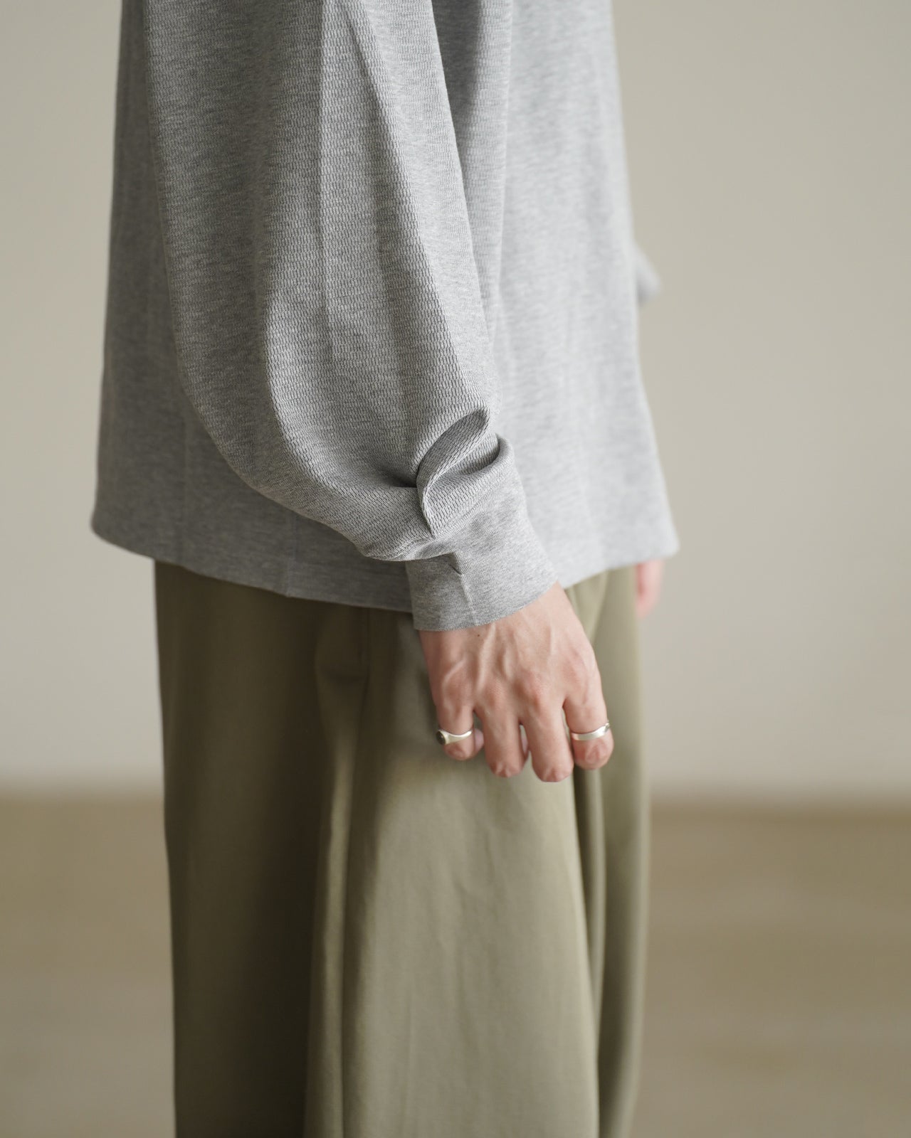 グレーの長袖Tシャツ本体を横から捉え、袖口のタックとリブの質感が際立つクローズアップ。裾はやや長めでナチュラルなドレープを生むデザイン。