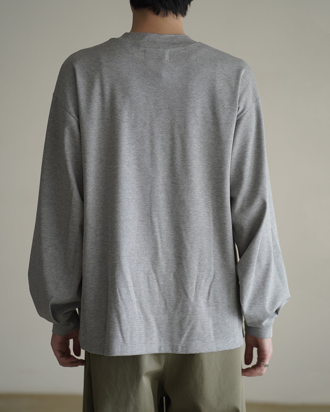 背面から見たライトグレーの長袖Tシャツ。生地は細かい編み柄が特徴で、裾までゆったりしたドロップシルエット。袖口はリブ仕様、首元は襟ぐりの浅めなクルーネック。