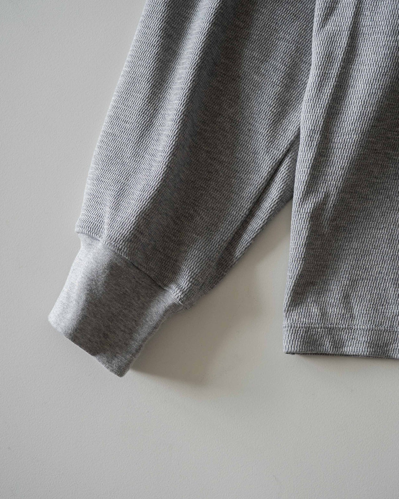 薄く光沢のあるサーマル素材の長袖Tシャツ。袖はプリーツ入りで立体感があり、布地の織り柄が細かな格子状に見える接写。色はライトグレー系。