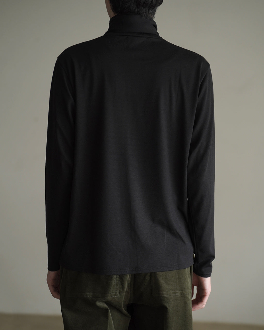 背面から見るWASHABLE-WOOL HIGH NECK SHIRT、黒系の長袖シャツ。裾がやや広がり、ネックラインが高めのハイネックデザインが印象的。