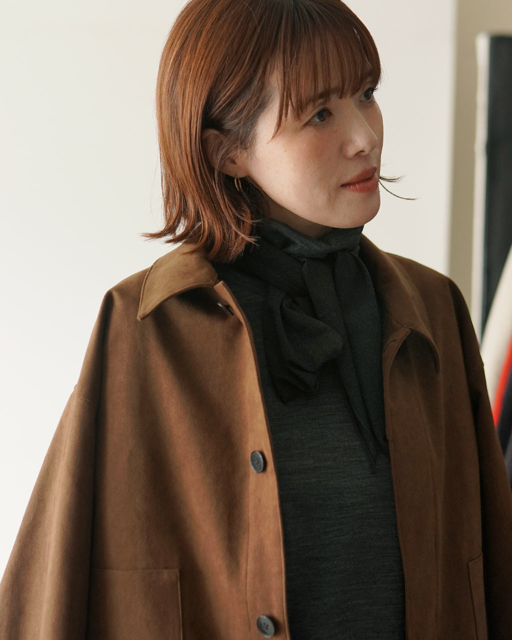 Wool混合の高ネックシャツを着用した女性の横顔アップ。焦げ茶色のボリュームあるコートに、黒いリボン風ネックが映え、落ち着いた雰囲気。