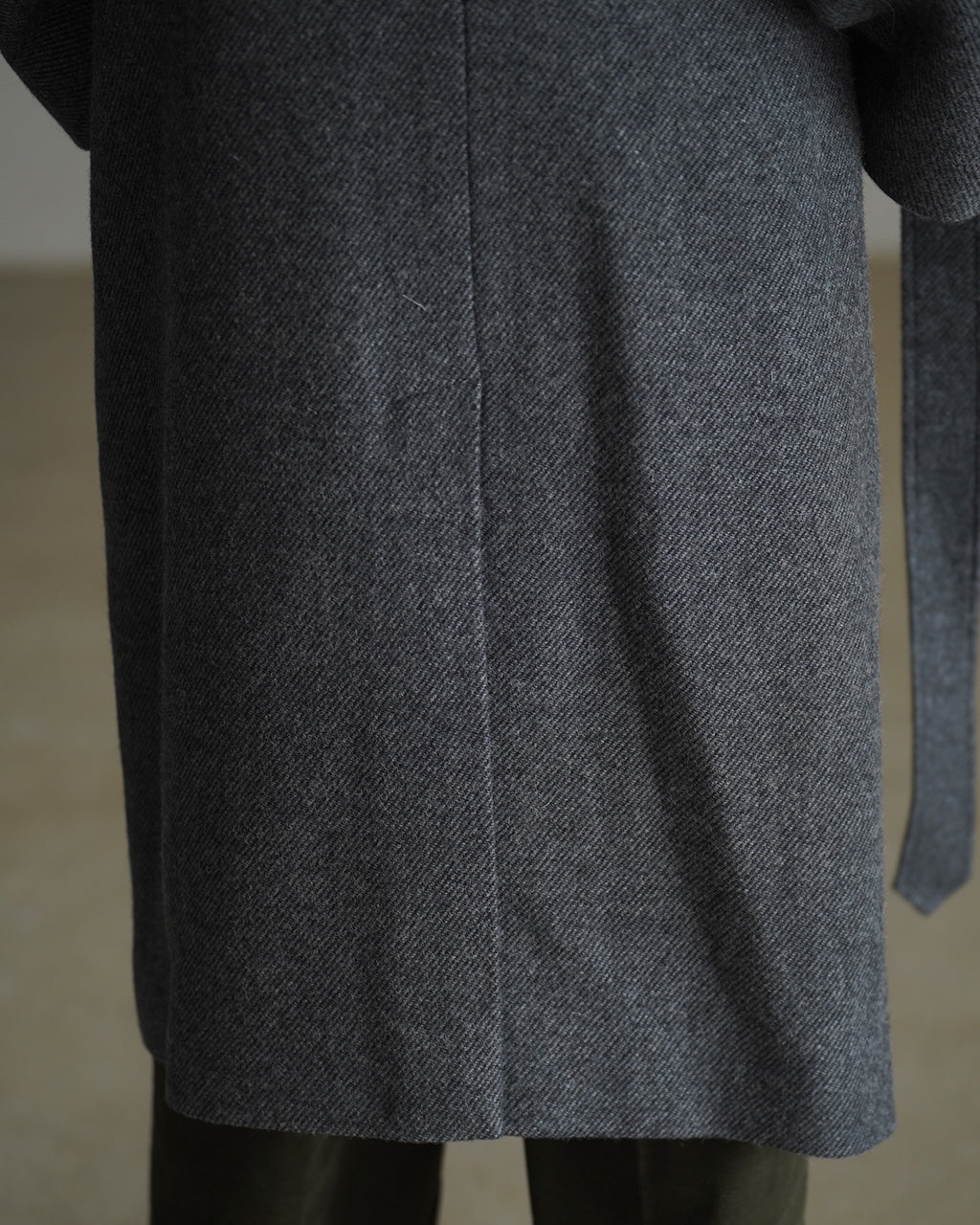濃いグレーのウール混リバーコートを裾付近から寄ったアップで撮影。生地の繊細な畝と縫製の美しさが分かるディテールショット。