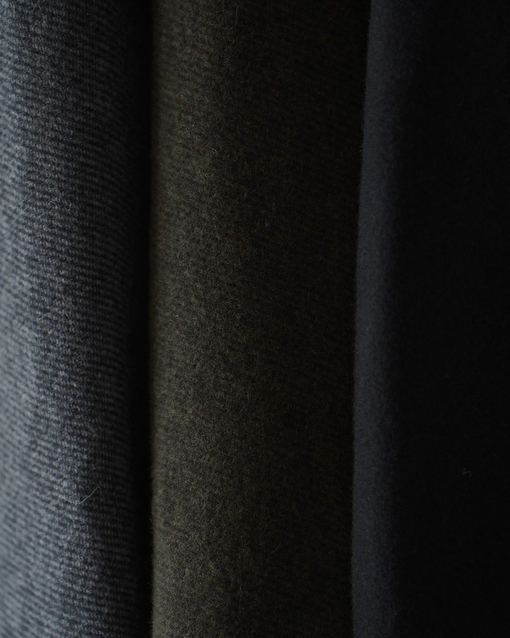 WRAP COAT RM262-034の生地切替が近接で見える黒系の織り素材。幅広い布量が重厚感とドレープを生み、左側から右へ連なる布地の陰影が分かるディテール写真