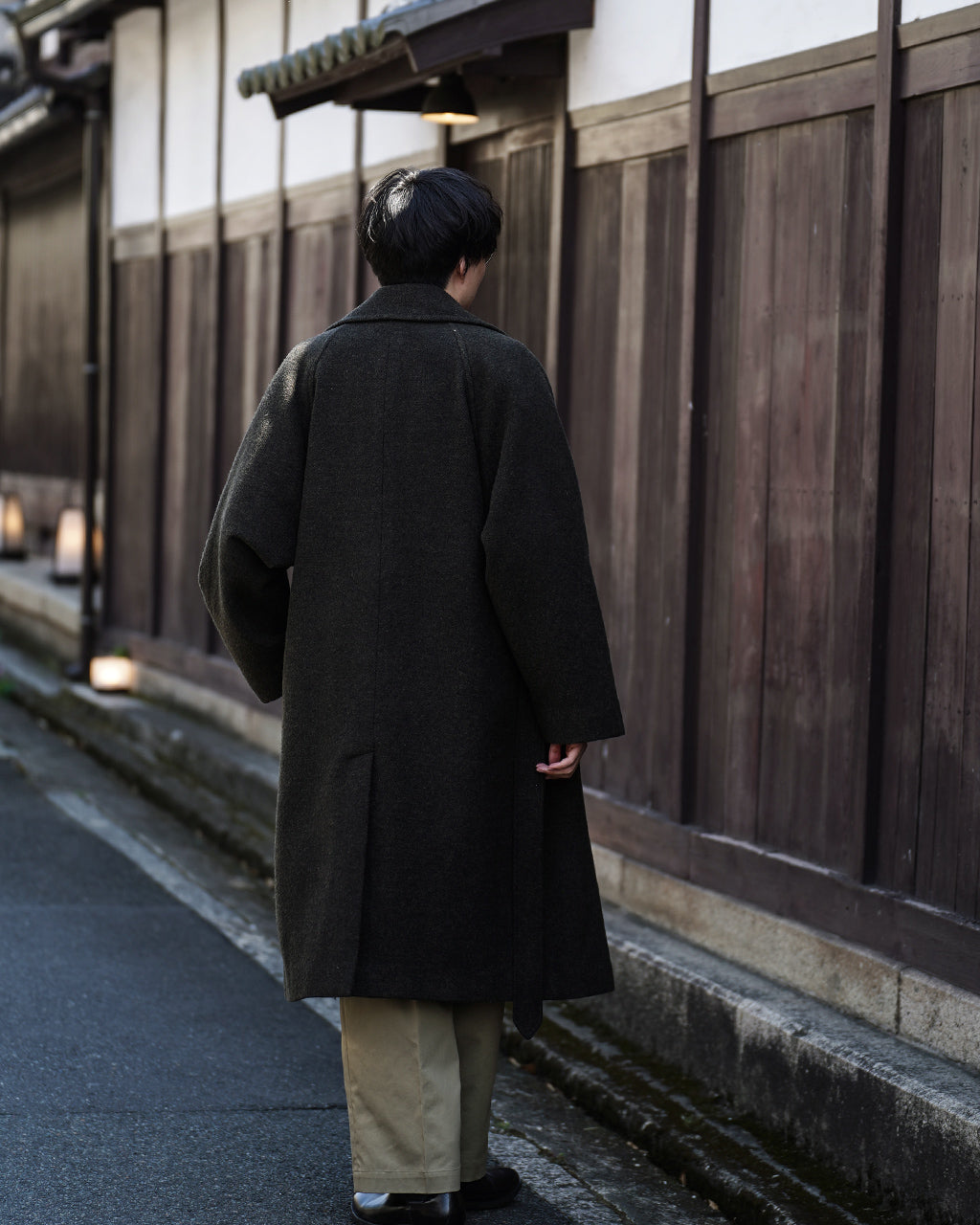 後方から撮影されたWRAP COAT RM262-034。深いグレーのコートが木製の壁の前に広がり、ゆったりとしたシルエットと袖口の形が見える。ベルトを締めず自然なドレープ感が伝わる背面ショット。