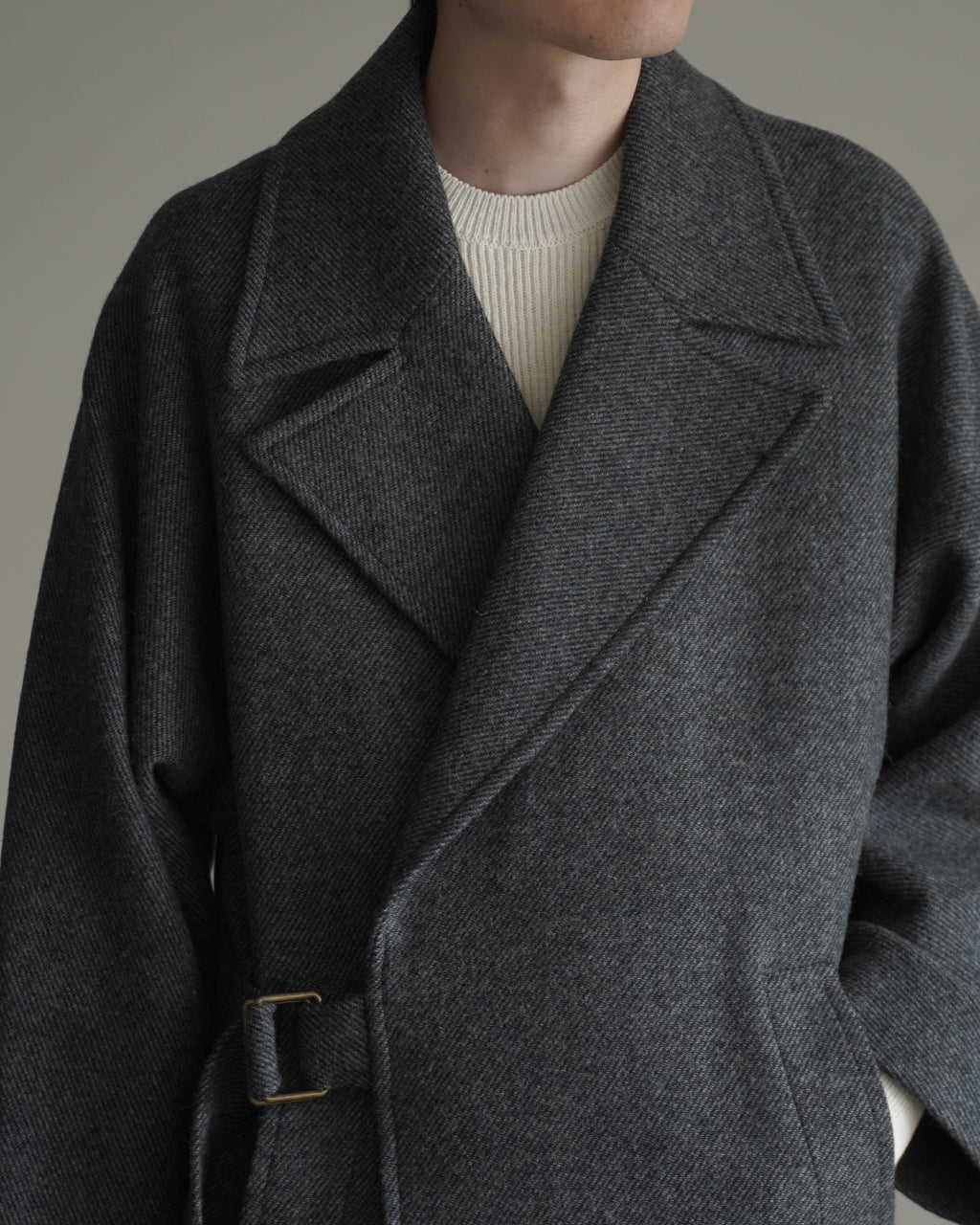 ダークグレーのウール混素材のWRAP COAT RM262-034のアップ。大きめの襟と前締まりのあるデザイン、肩から袖へ続く一枚布のパターンが特徴的。ベルト留め金具が控えめに見える正面ショット。