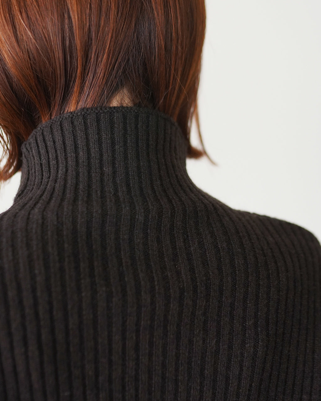 黒のリブニットの後ろ姿。高めのタートルネックと立体的なリブ編みが特徴で、肩付近の縦リブが強調されている様子。