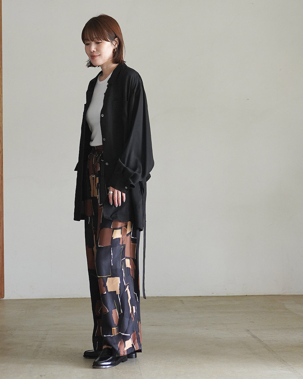 SACRA Geometric Painting Pantsのモデル着用カット。黒の長袖シャツと幾何学柄パンツを組み合わせ、ゆったりとしたワイドシルエットと落ち着いた色味が印象的
