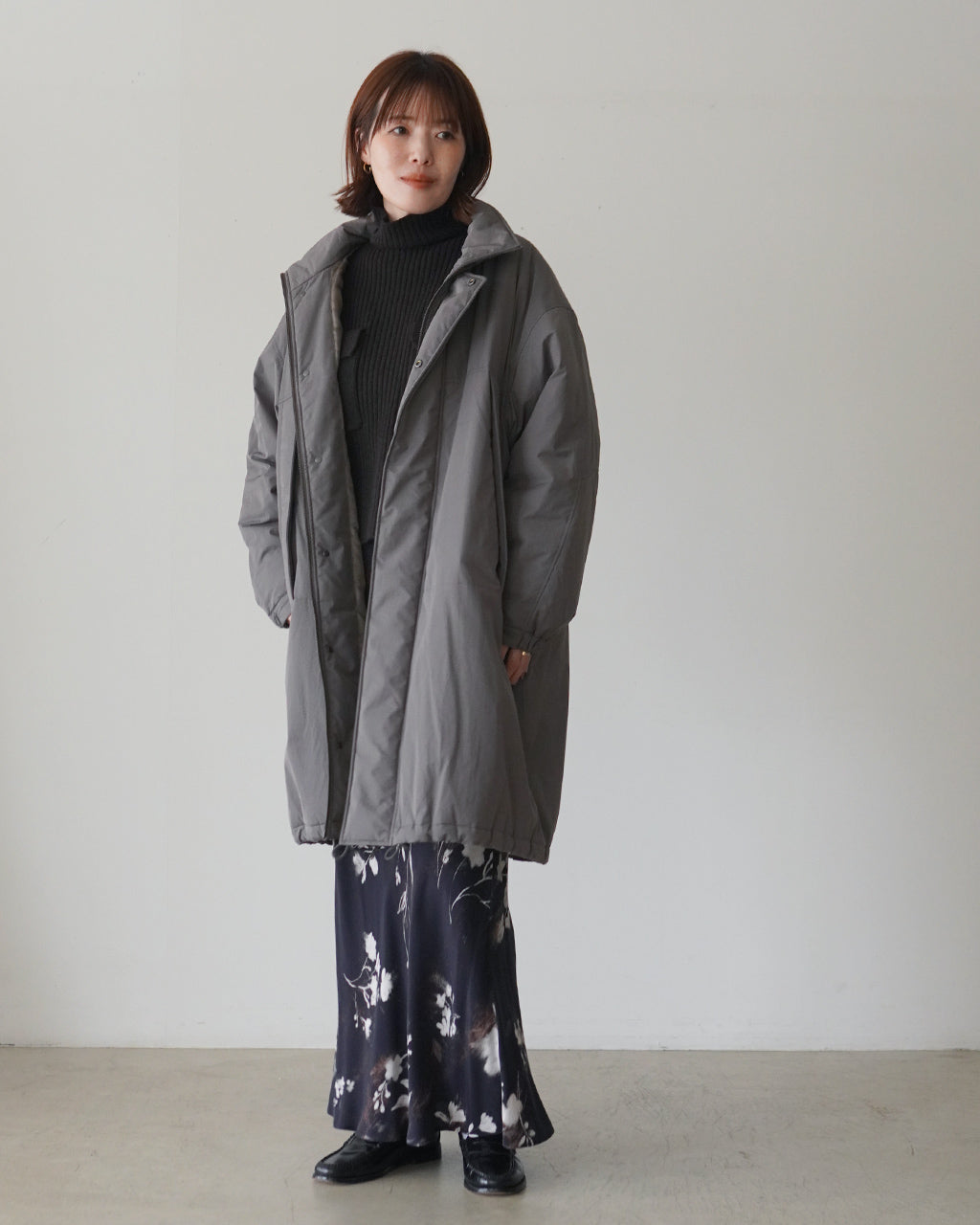 カーキの中綿コートを着用した女性モデルの正面姿。膝下丈でゆったりとしたシルエット、マットなタフタ地と内側のキュプラ裏地が見える。前開きはジップとボタンで開閉可能、裾はやや広がらずすっきり見える。
