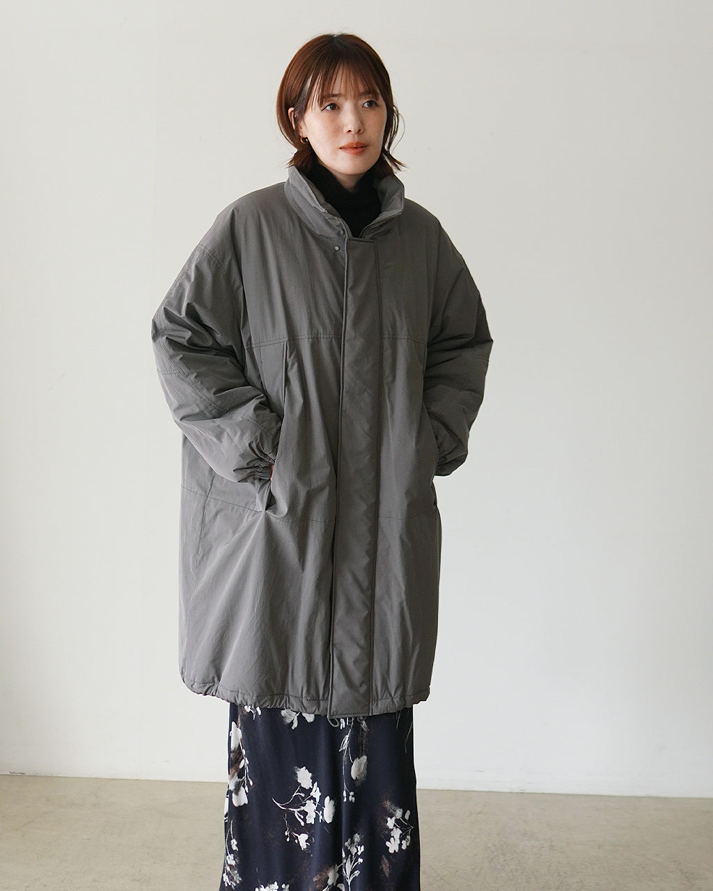 SACRAのカーキ系中綿コートを着用した女性モデルの前方ショット。ゆったりとしたシルエットで膝下まで覆う長め丈、立ち上がったスタンドカラーとジップ開閉デザインが特徴的。
