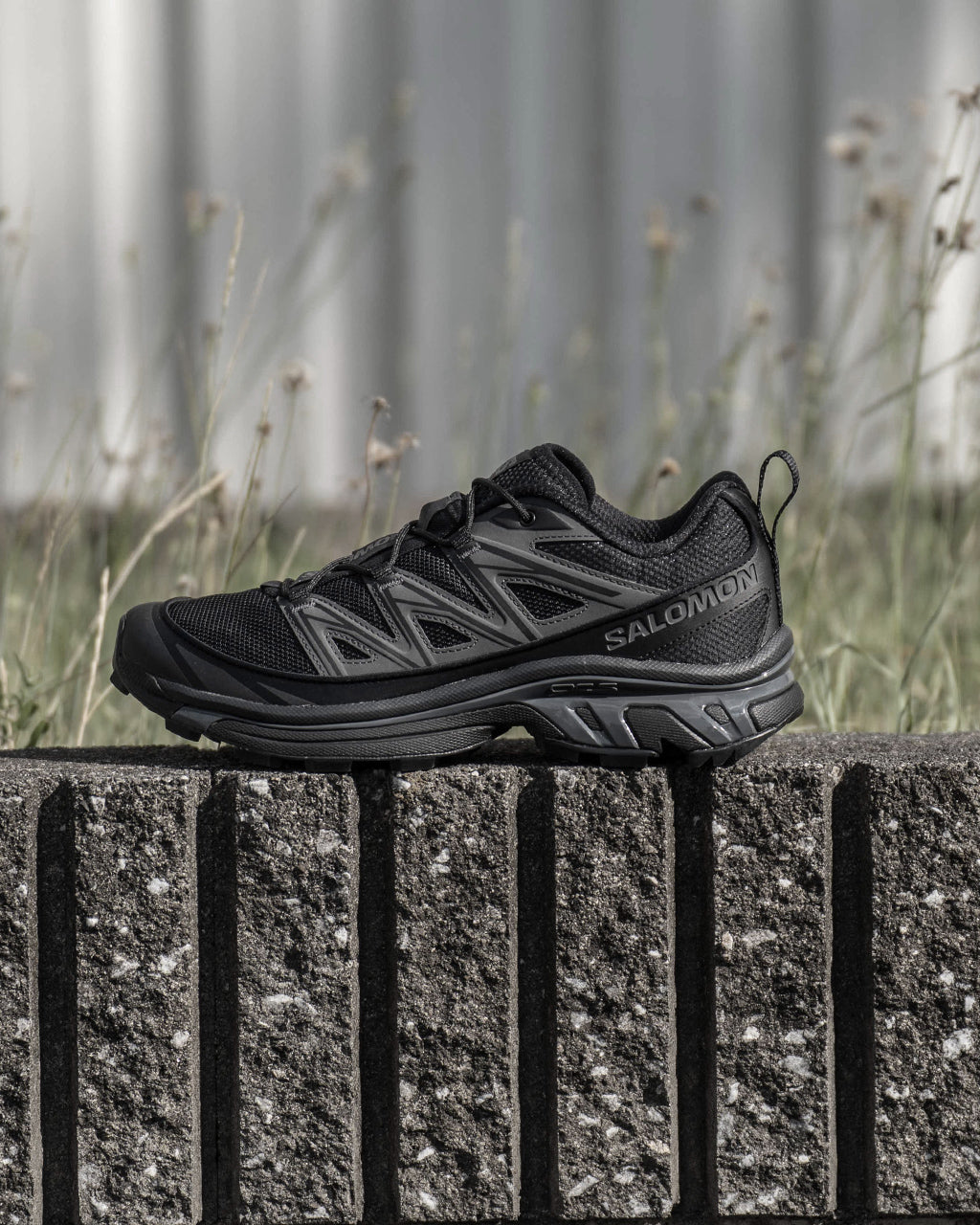 黒系のSALOMON XT-6 EXPANSEが地面のコンクリートブロックの上に置かれ、サイドから見る全体像。通気性のあるメッシュと立体的なアッパー、ブランドロゴが際立つデザインです。
